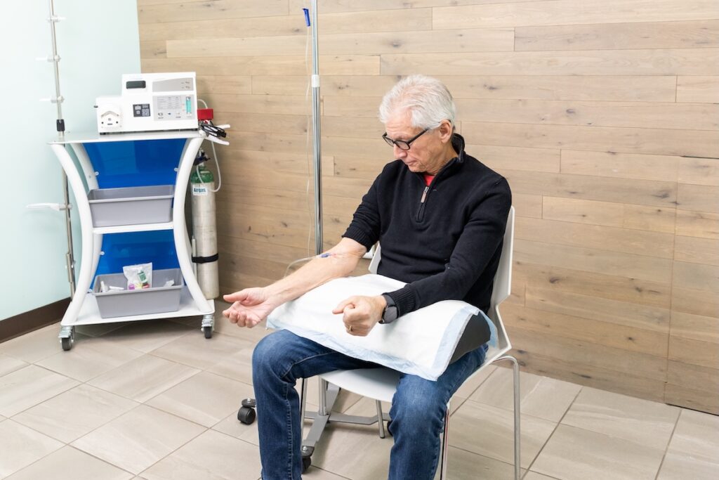 Man sits in chair of medical office with pillow resting on his knees and both arms placed on pillow. Attached to his arm is a needle connecting him to an infusion of IV hydration in North Liberty.