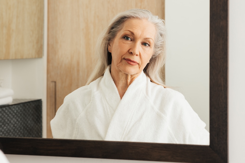 Woman looking in the mirror while wearing a white robe, wondering how to age gracefully with treatment for skin tightening in North Liberty