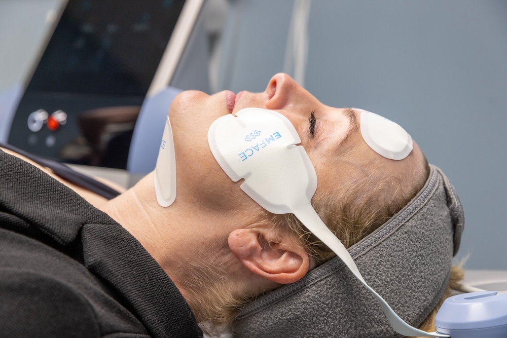 Woman lays down and relaxes with eyes closed with applicators placed on her cheeks to contract muscles and heat deep skin layers for her skin tightening in North Liberty