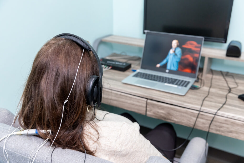 Woman wears headphones and watches a video on a laptop while connected to a neurofeedback machine which helps her modify her thought patterns. This treatment was developed after measuring her brainwave activity using brain mapping near Iowa City.