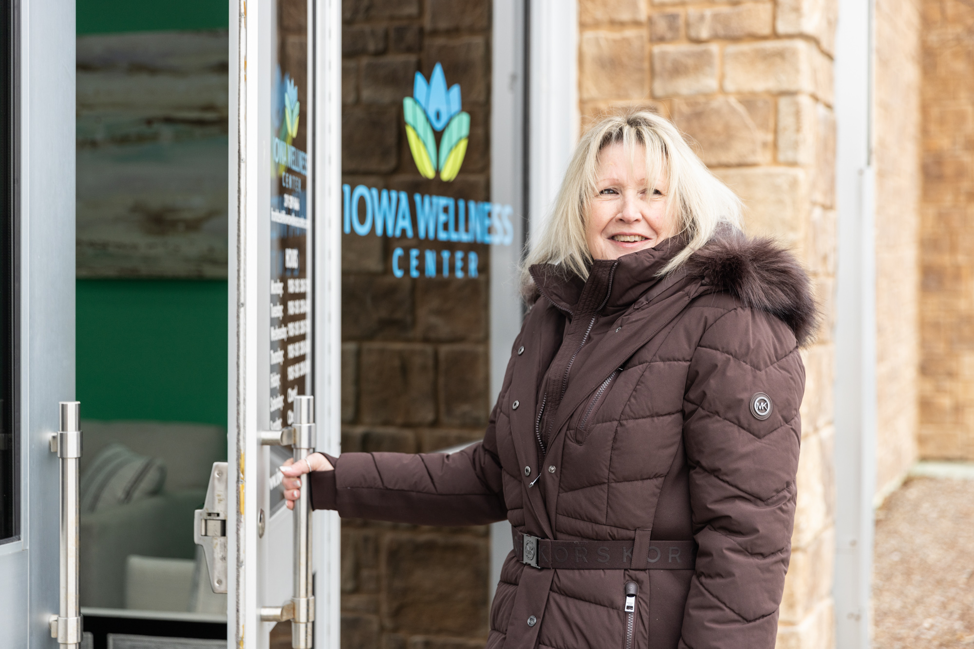 Patient walking into a weight loss center near Cedar Rapids. She is wearing a winter coat with fur on the hood and is opening the door. Beside her, a sign displays the name of the clinic, "Iowa Wellness Center."