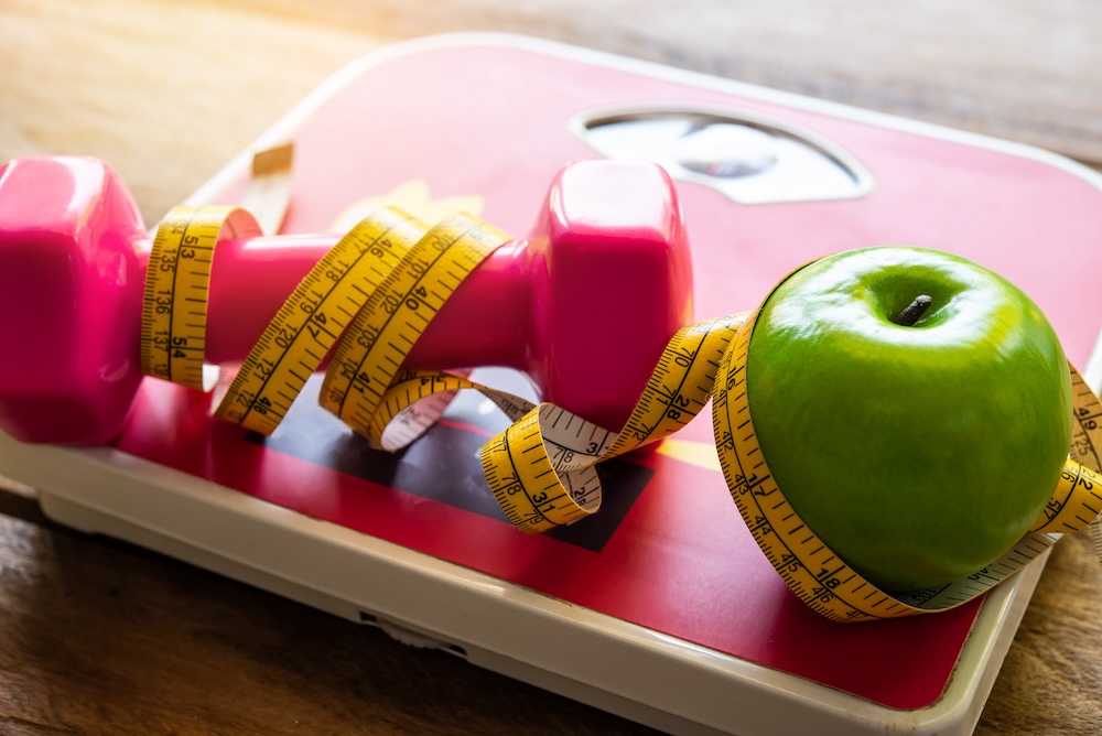 Photo a scale, pink weight, and a green apple with measuring tape wrapped around them because they are essential to treatments like weight loss injectables in North Liberty